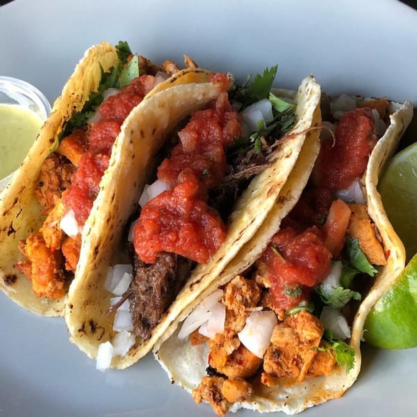 Three tacos with various fillings, salsa, onions, cilantro, and a lime wedge on a white plate.
