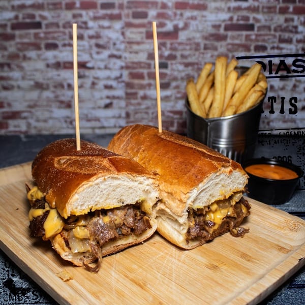 Cheesesteak sandwich cut in half with fries and dipping sauce on a wooden board.