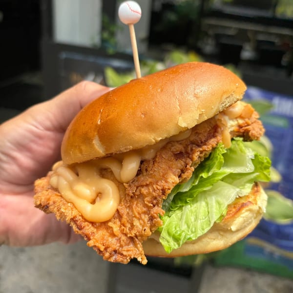 Hand holding a Hangar 11 fried chicken sandwich with lettuce, sauce, and a baseball toothpick.