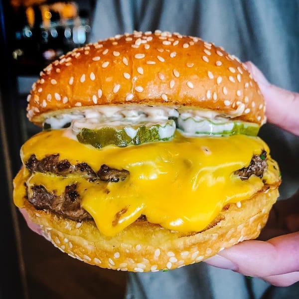 A hand holds a Hangar 11 double cheeseburger with pickles and special sauce on a sesame bun.