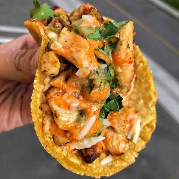 Hand holding a crispy taco filled with chicken, orange sauce, green salsa, and cilantro.