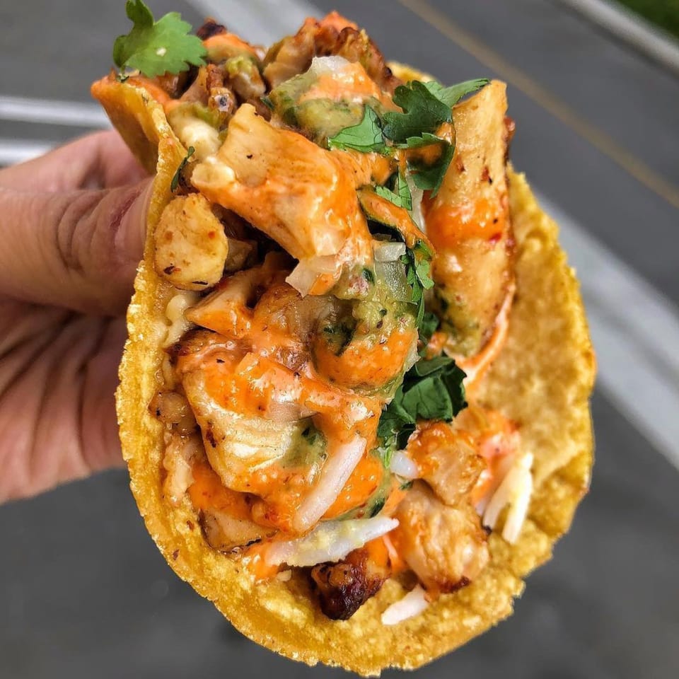 Hand holding a crispy taco filled with chicken, orange sauce, green salsa, and cilantro.