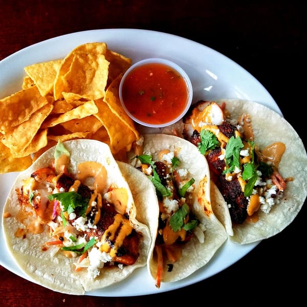 Three fish tacos with cilantro, cheese, and sauce, served with tortilla chips and salsa.